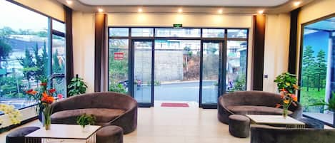 A modern lobby with plush seating, a chandelier, and large windows offering a view of the street and greenery outside.