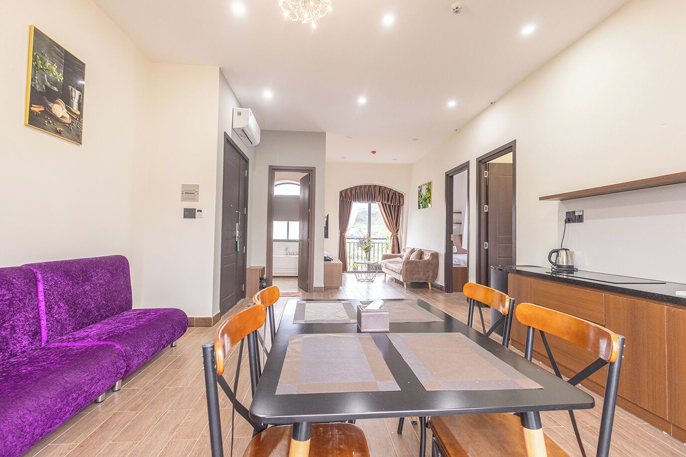 A modern hotel room with a dining area, a purple sofa, a kitchen, and a balcony with a view.