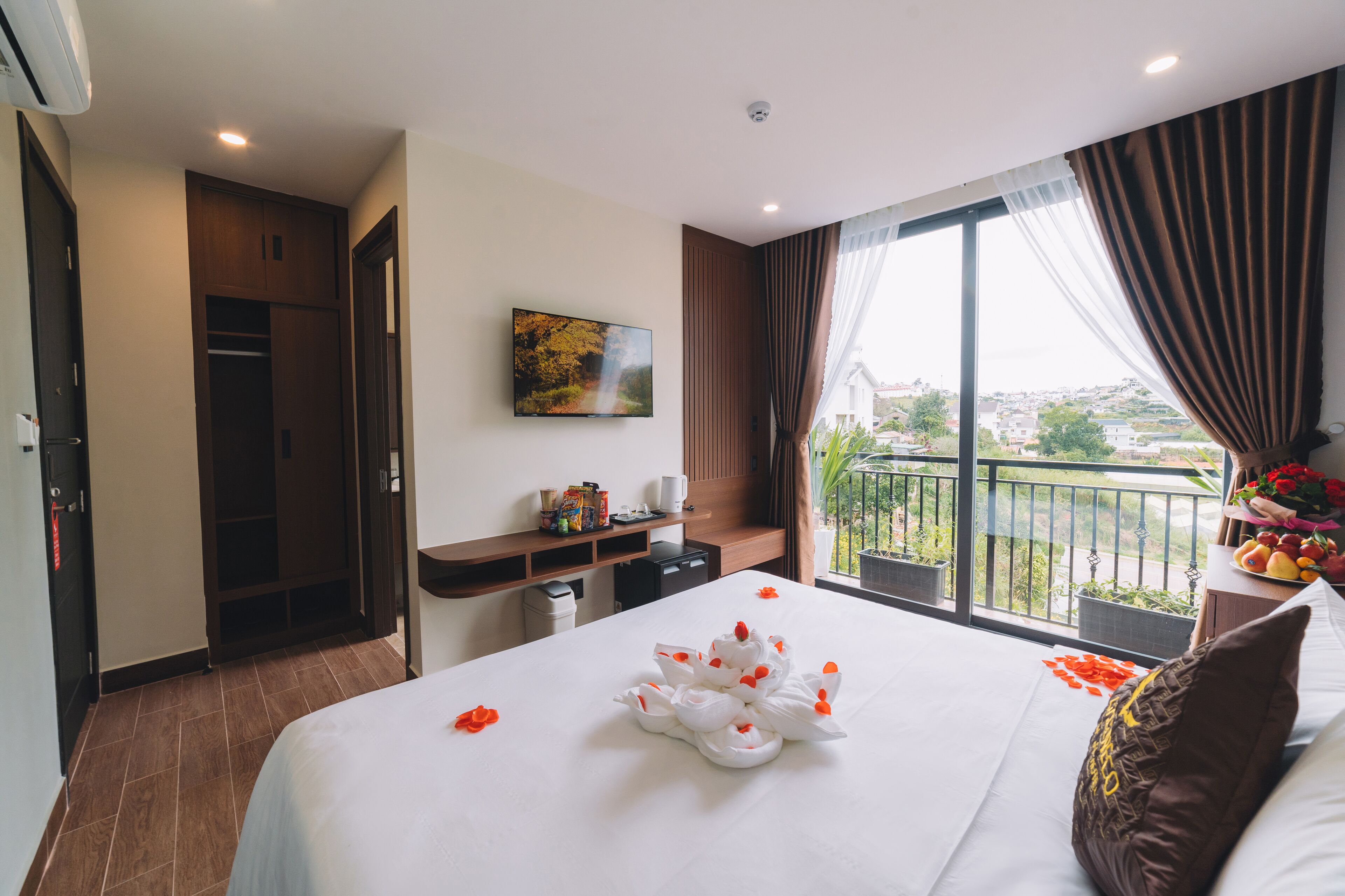 A hotel room with a large bed, a desk with a computer, a small seating area, and a balcony with a view.