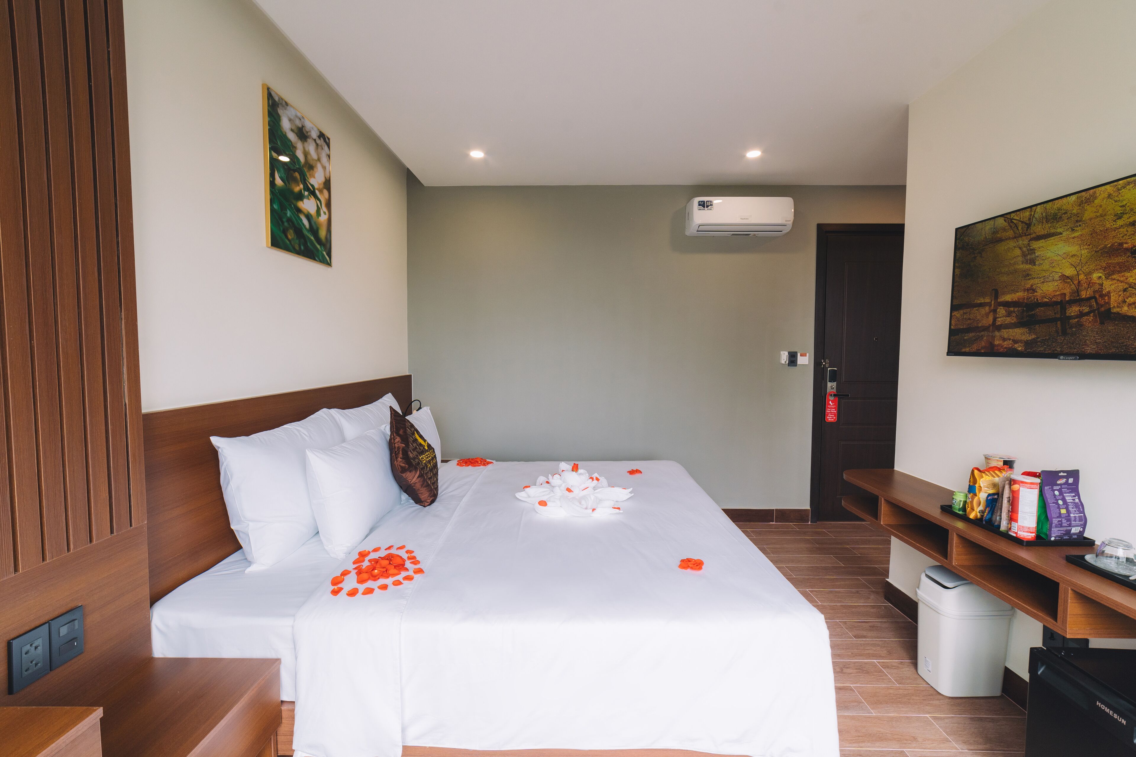 A hotel room with a neatly made bed, a wooden headboard, a wall-mounted air conditioner, a painting, and a shelf with snacks and a tray.