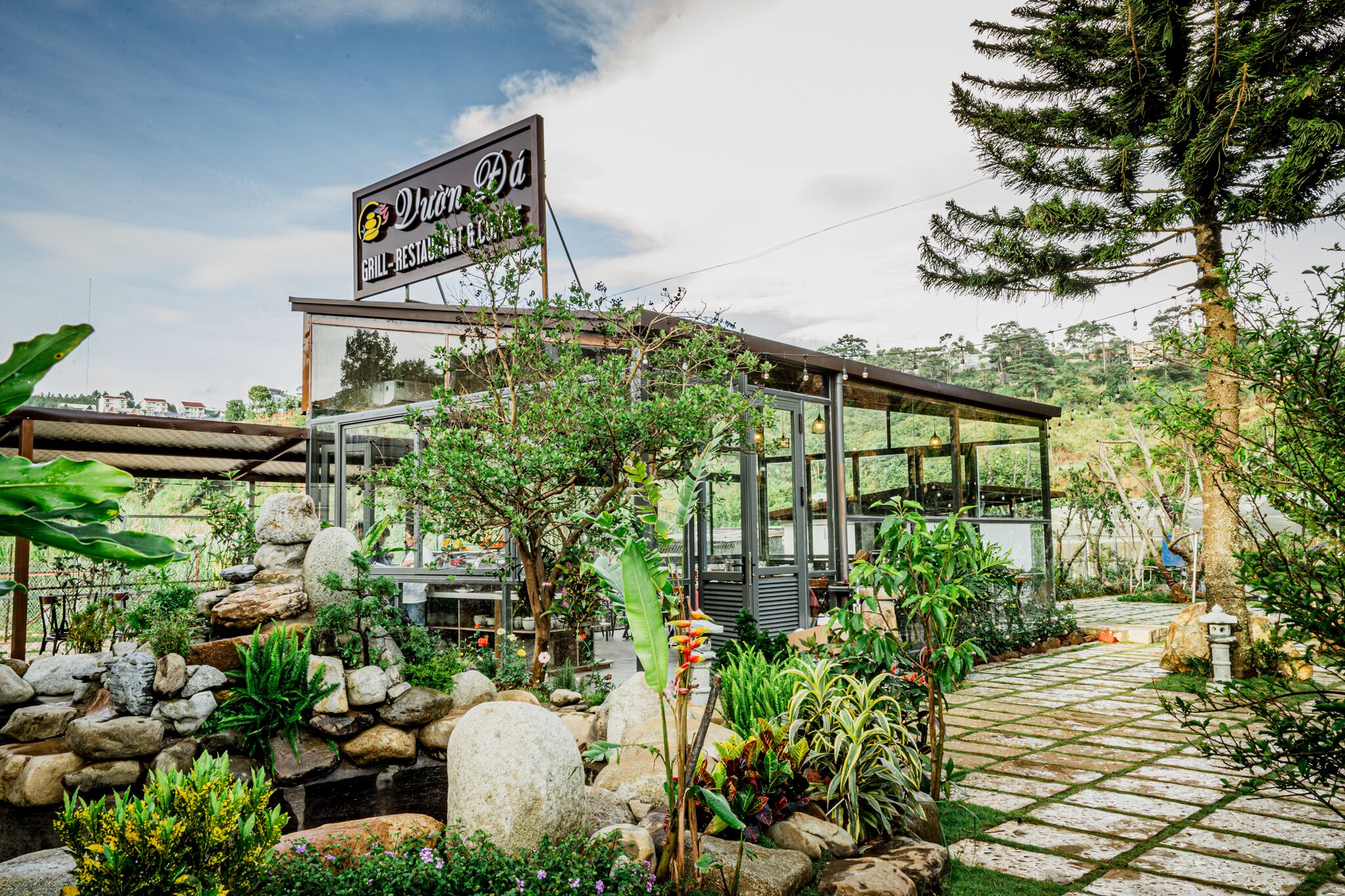 Un restaurant doté d’une structure en verre, entouré d’un jardin agrémenté de rochers et de plantes.
