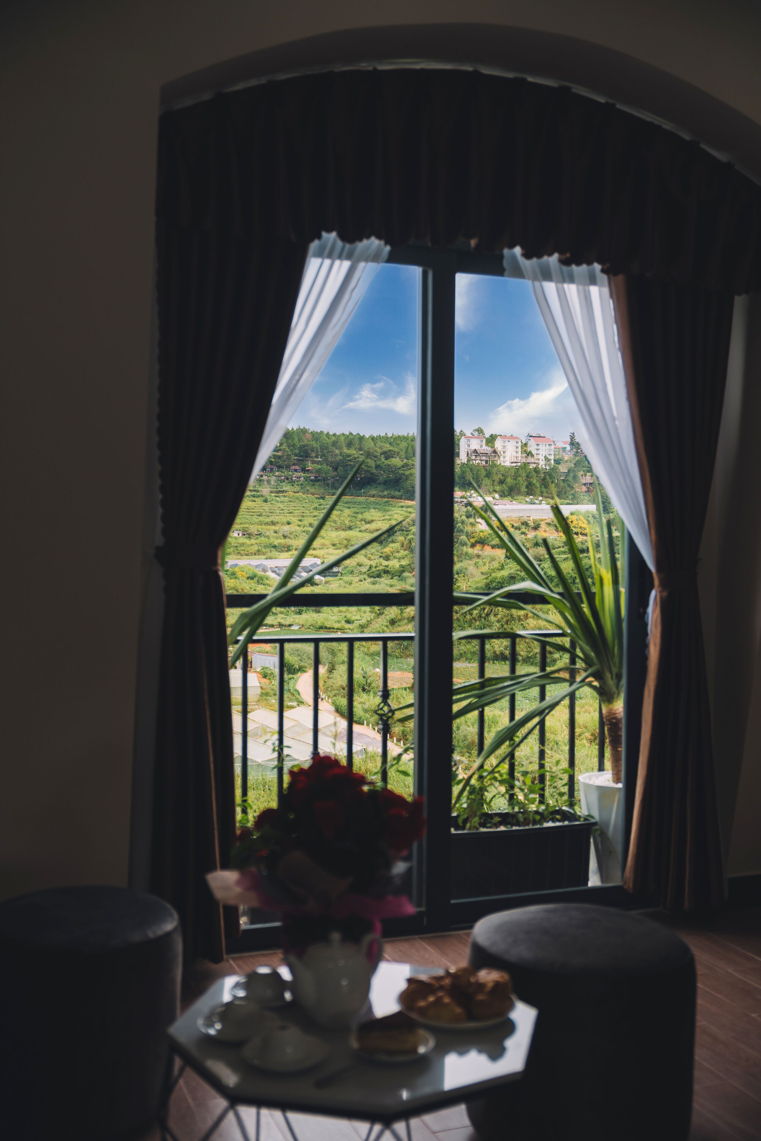 Une pièce avec un balcon donnant sur un paysage verdoyant et des bâtiments. On y trouve une table avec un vase de fleurs et une assiette de pâtisseries.