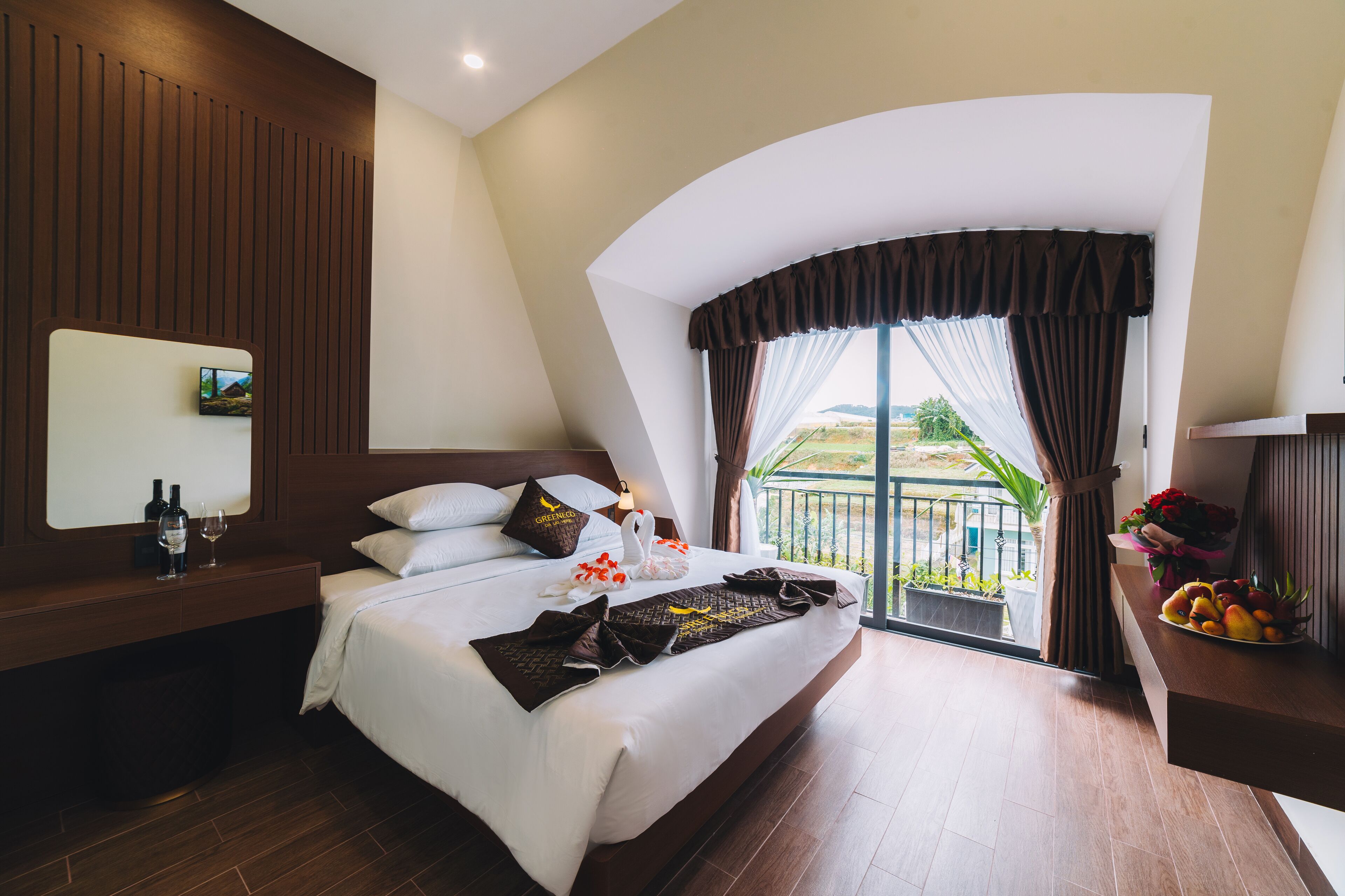 A modern hotel room with a large bed, a wooden headboard, a bedside table with a mirror, and a view of the outdoors through a large window.