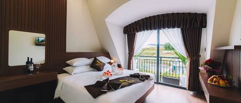 A modern hotel room with a large bed, a wooden headboard, a bedside table with a mirror, and a view of the outdoors through a large window.
