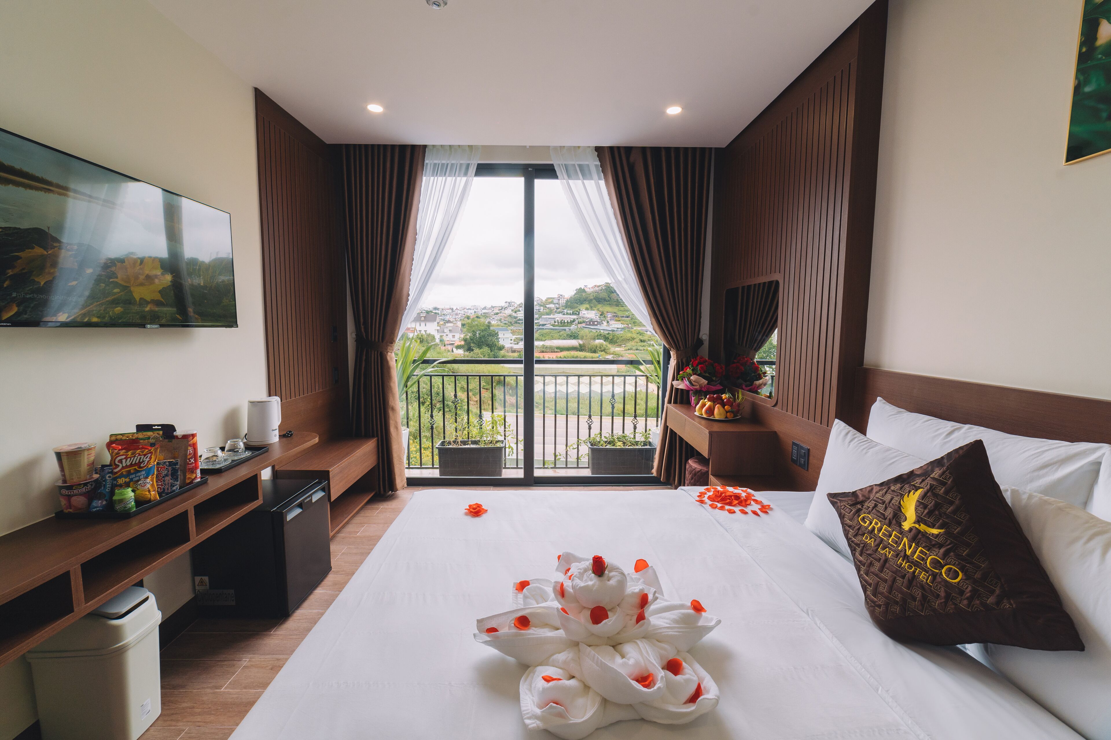 A hotel room with a bed, a TV, a balcony with potted plants, and a minibar.