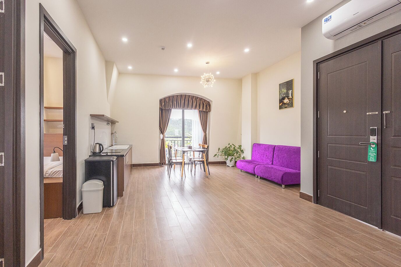 A modern hotel room with a wooden floor, a purple sofa, a dining area with a table and chairs, and a large dark door.