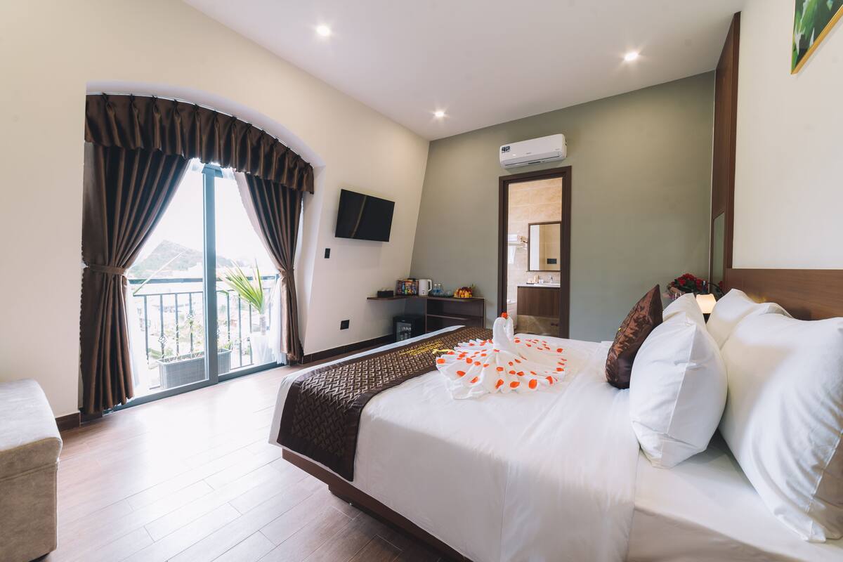 A hotel room with a large bed, a TV mounted on the wall, a balcony with curtains, and an air conditioning unit.