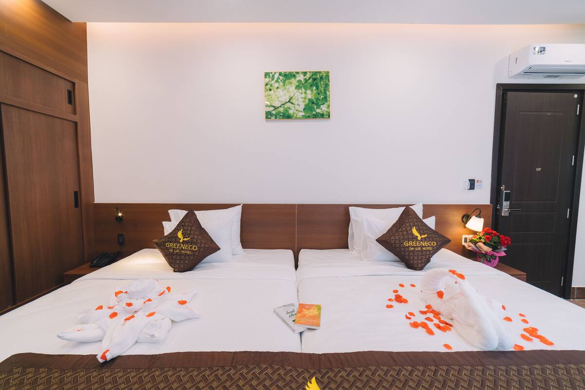 A hotel room with a double bed, two pillows with the hotel logo, a bedside table with a lamp and flowers, and a wall-mounted air conditioner.