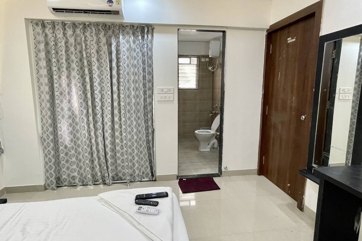 Une chambre d’hôtel avec un lit, une salle de bain visible à travers une porte ouverte, un miroir et un climatiseur fixé au mur.