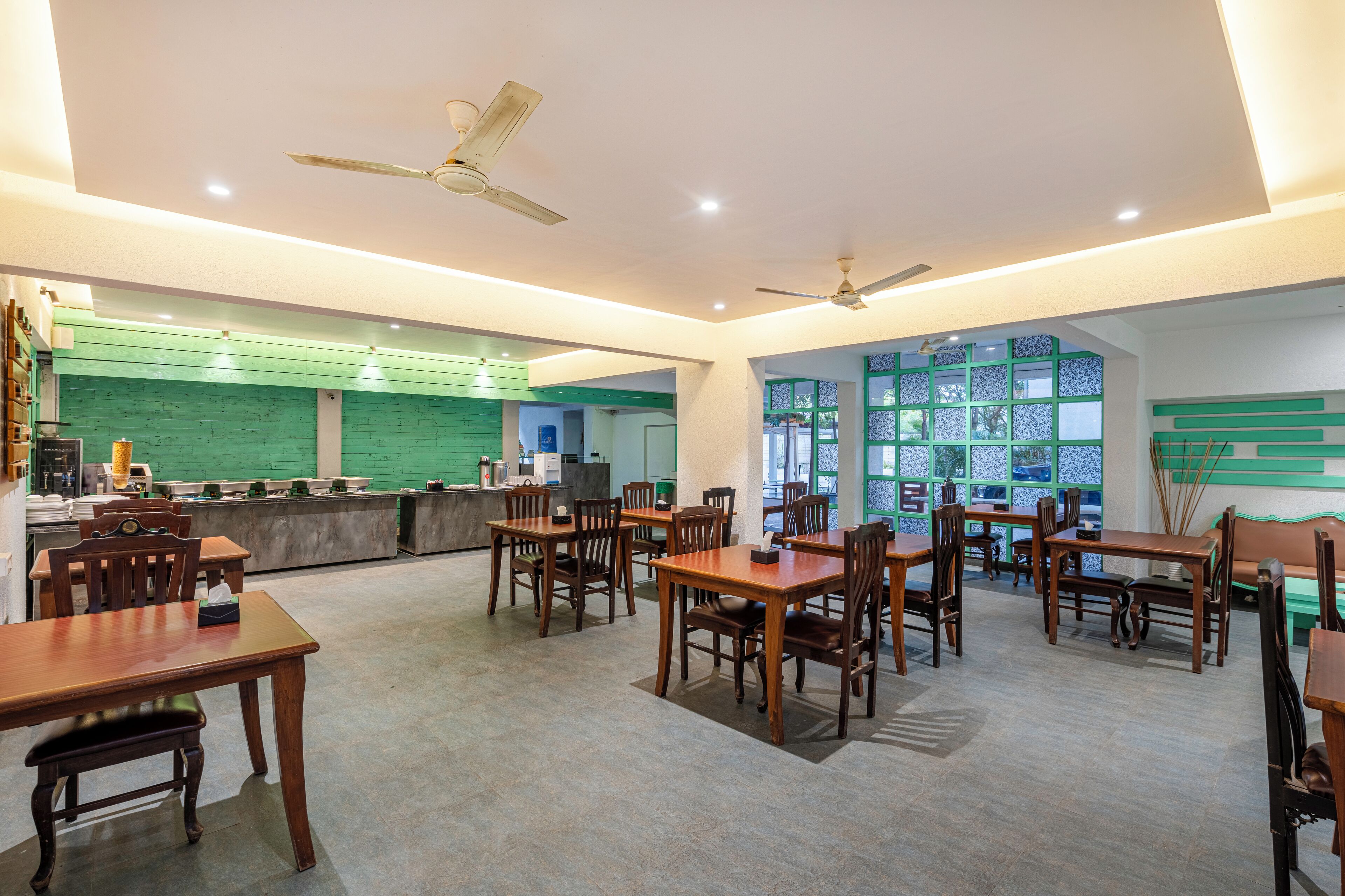 A spacious dining area with multiple wooden tables and chairs, a counter with a green backdrop, and ceiling fans.