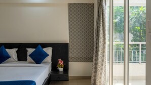 A hotel room with a bed, bedside table, air conditioning unit, window with curtains, and a view of greenery outside.