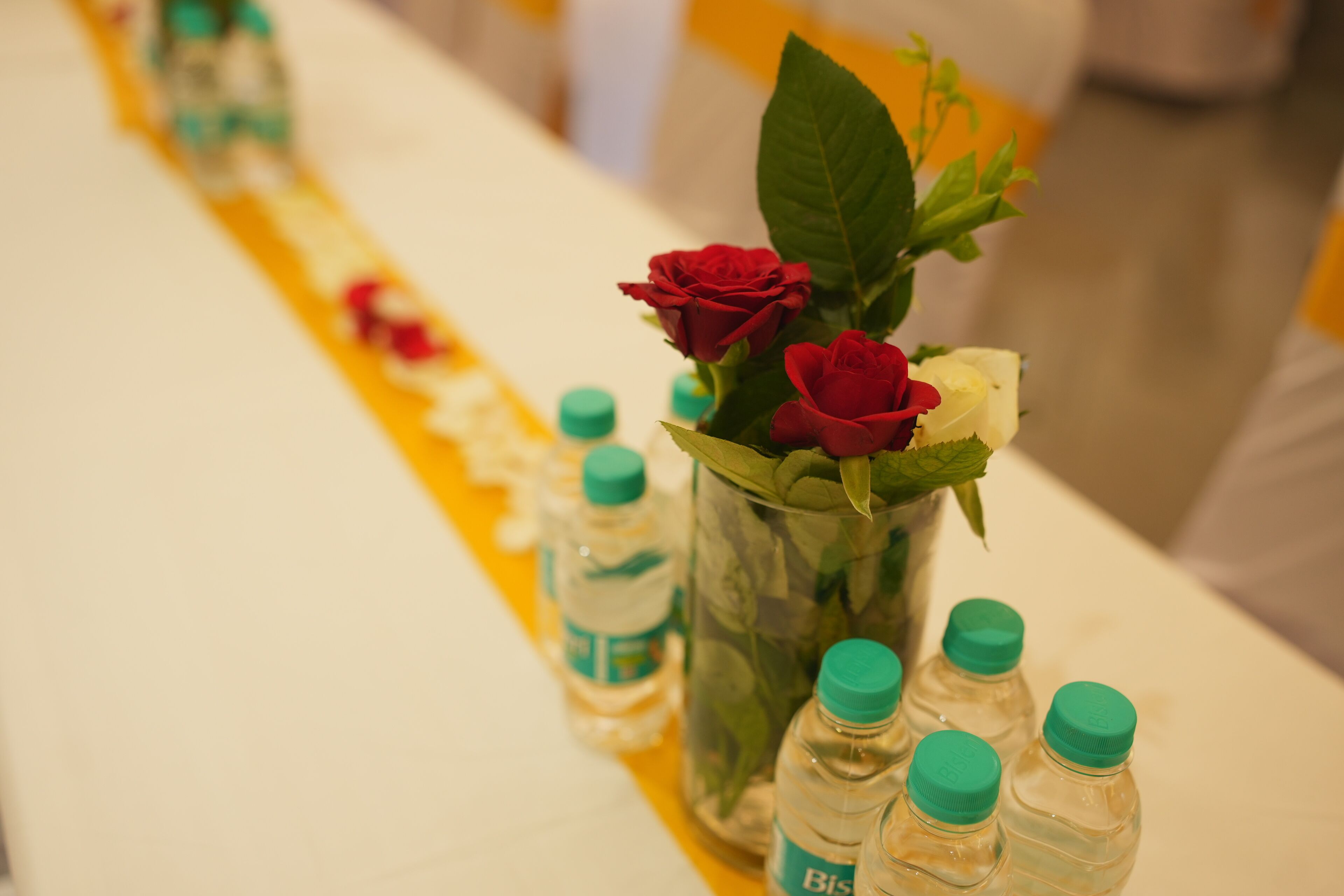 Una mesa con un centro de flores, botellas de agua y un camino de mesa amarillo.