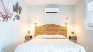 Un dormitorio con cabecera trenzada, colcha blanca acolchada, dos lámparas de noche y aire acondicionado instalado en la pared.
