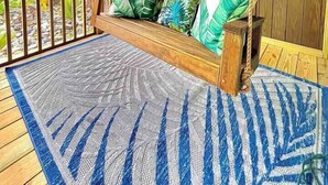 Una terraza de madera con una mecedora, un tapete estampado y vistas a edificios y palmeras.