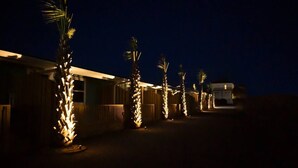 Eine Reihe von Palmen mit Lichterketten, ein Gebäude mit Satteldach und ein Fahrzeug im Hintergrund.
