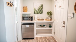 A compact kitchen with a microwave, coffee maker, and a refrigerator. There is a shelf with decorative items and a door with a 'No Smoking' sign.