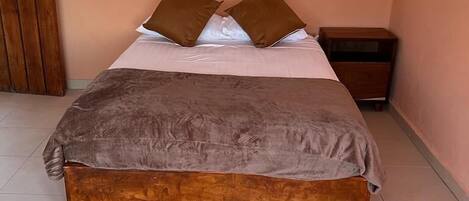 A bedroom with a large bed, two pillows, a wooden nightstand, and a window with curtains.