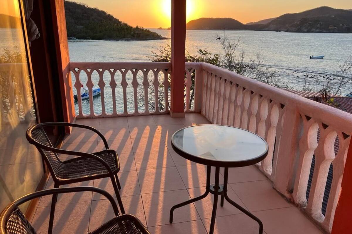 Un balcón con dos sillas de mimbre y una mesa de vidrio, con vista a un cuerpo de agua donde se ve una lancha y colinas lejanas durante el atardecer.