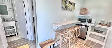A compact kitchen with a white countertop, a microwave, and a coffee maker. There are three wooden stools and a bench.