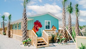 Ein Strandhaus mit einem Hahn-Wandbild, einer Holzterrasse und Palmen.