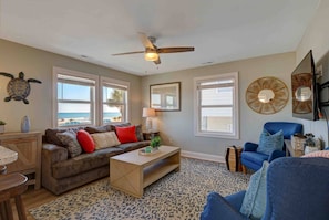 A living room with a sofa, coffee table, and blue armchair, large windows with ocean views, and a ceiling fan.