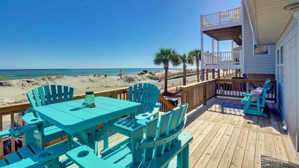 Eine Holzterrasse mit türkisfarbenen Stühlen und einem Tisch, mit Blick auf einen Strand mit Palmen und einem klaren Himmel.