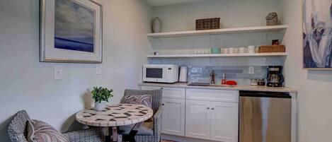A compact kitchen with white cabinets, a microwave, a coffee maker, and a dishwasher. There is a small round table with chairs and a potted plant.