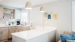 A modern kitchen with white cabinetry, a large island, pendant lights, and a view of the living area. - The Hideaway B- Beach - Dog Friendly - Playground - Bikes - Fenced in Yard (Surf City)