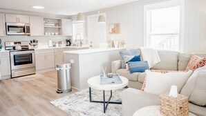 A modern kitchen with white cabinets, a microwave, oven, and a sink. There is a dining area with a white table and chairs, and a living room with a beige sofa and decorative pillows. - The Hideaway B- Beach - Dog Friendly - Playground - Bikes - Fenced in Yard (Surf City)