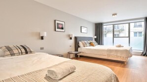 Una habitación de hotel moderna con dos camas, una mesita auxiliar pequeña, un ventanal grande con cortinas y un televisor.
