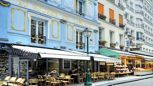 Calle con cafés y tiendas, incluyendo un edificio azul con el rótulo 'AU ROCHER DE CANCALE'.