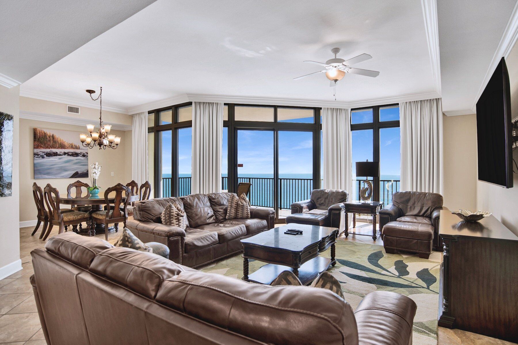 A spacious living room with leather sofas, a coffee table, and a dining area with a chandelier. Large windows offer a view of the ocean.