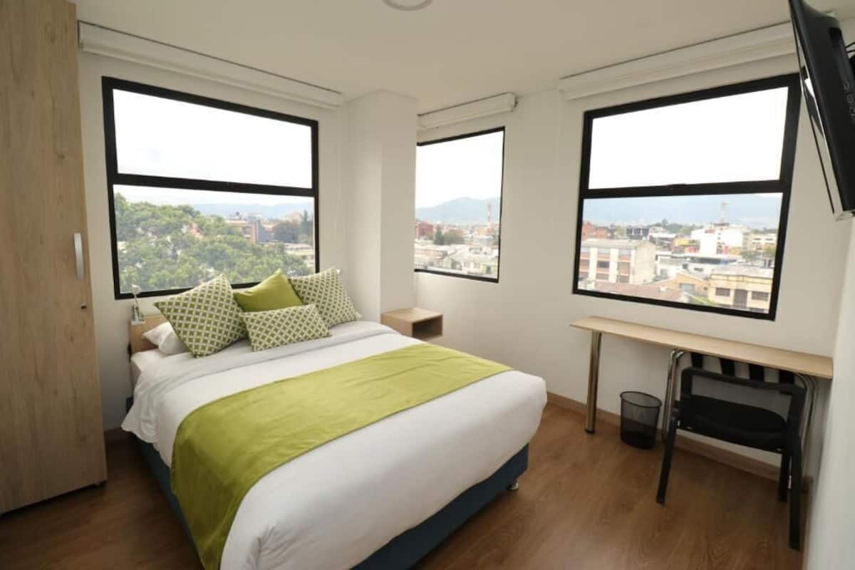 A bedroom with a bed, two windows, a desk, and a chair.