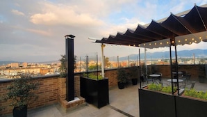 En tagterrasse med udsigt over bybilledet, med en pergola med lyskæder, plantekasser og udendørs møbler.