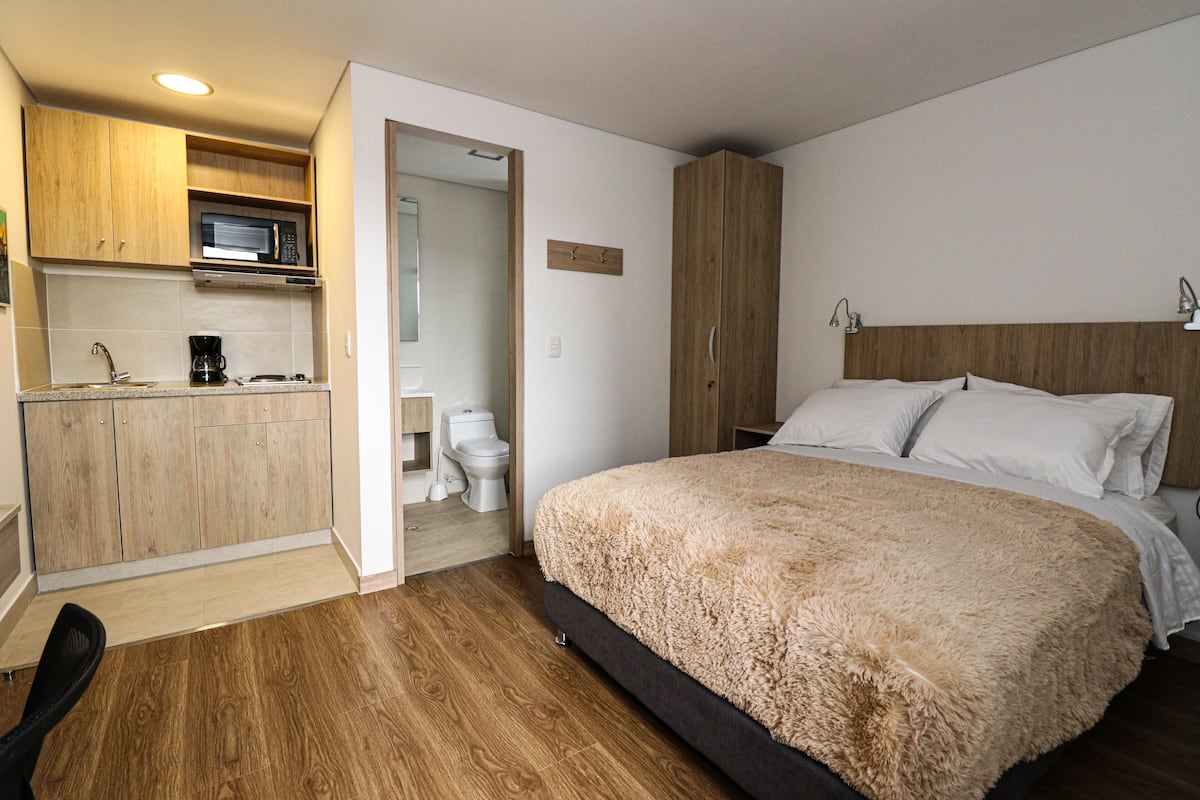 A bedroom with a bed, wooden wardrobe, and a kitchenette with a microwave.