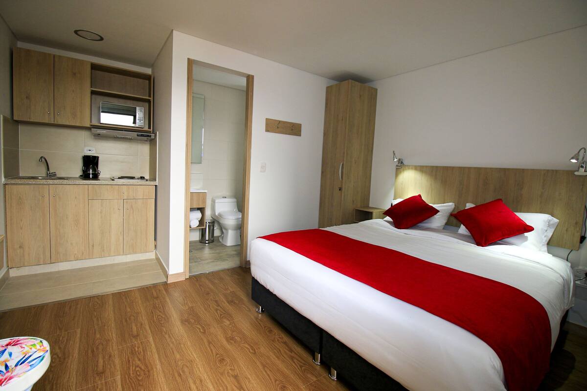 A hotel room with a bed, a red throw, a wooden headboard, a nightstand, a lamp, a wooden wardrobe, a microwave, a sink, and a toilet.