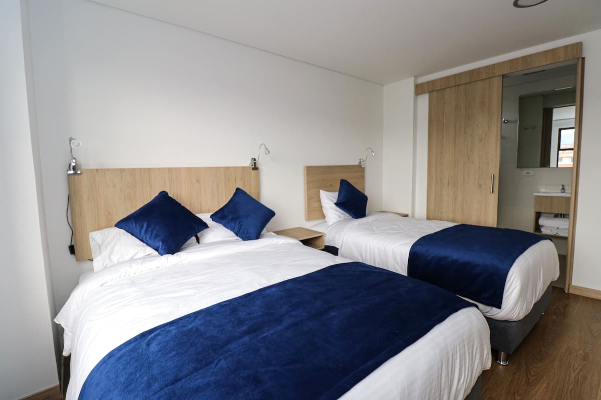 A hotel room with two beds, each with a wooden headboard, white bedding, and blue accents. There is a wooden wardrobe and a mirror on the wall.