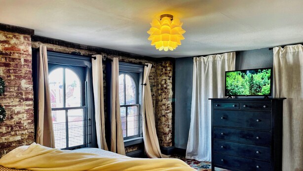 Um quarto com cama, cômoda, televisão e duas janelas com cortinas.