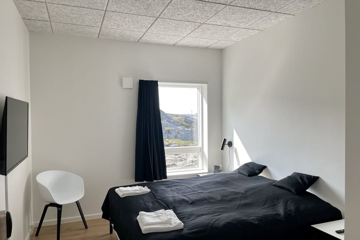 Une chambre d’hôtel moderne avec un grand lit, une chaise blanche, un bureau et une fenêtre donnant sur les montagnes.