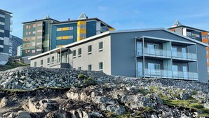 A modern multi-story building with a flat roof and large windows, situated on a rocky terrain with a clear sky. - Tuukkaq Apartments (Nuuk)