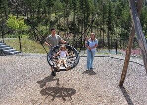 A playground with a swing set, a fenced area, and a forested hill in the background.