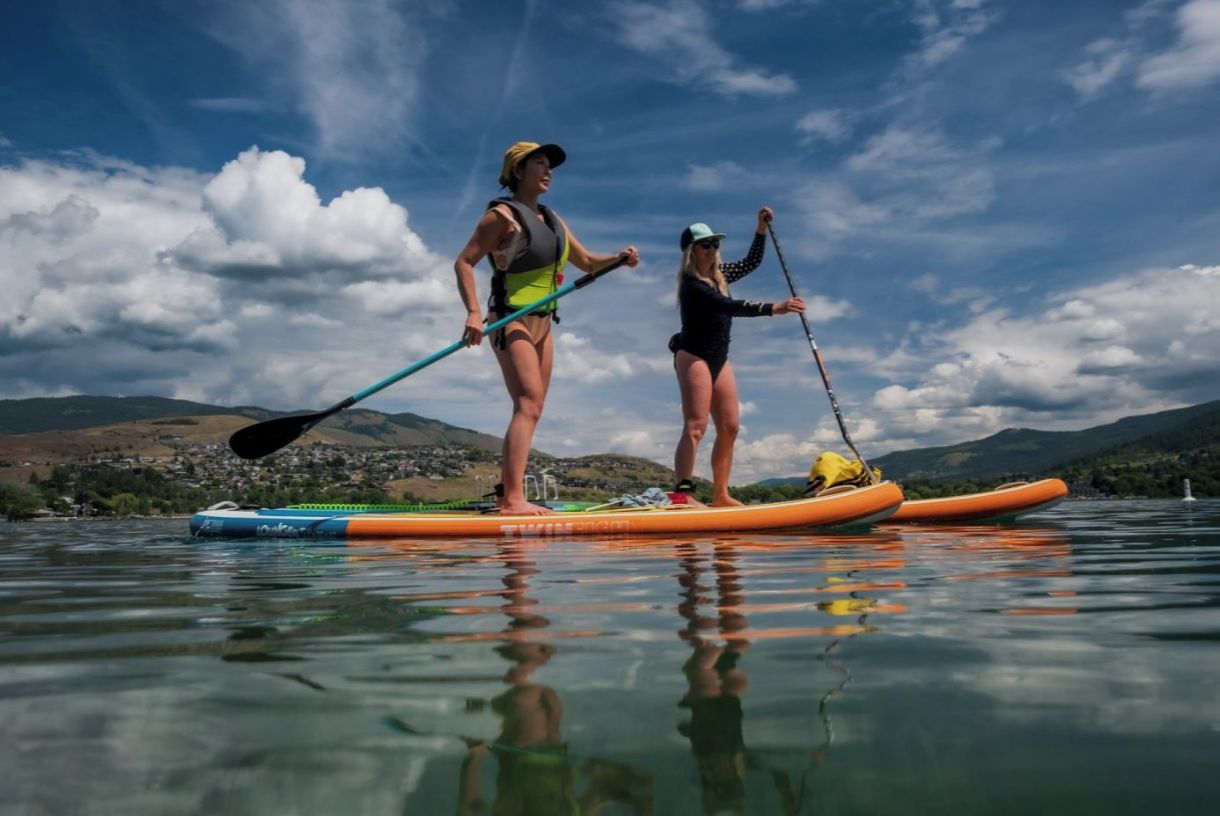 Two people paddleboarding on a calm lake with mountains in the background.