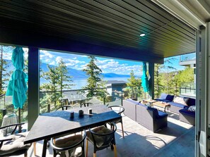 Een overdekt terras met een tafel en stoelen, met uitzicht op een waterpartij en bergen.