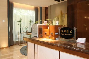 A hotel reception desk with a marble countertop, a visitor registration sign, a small plant, and a wall-mounted clock.