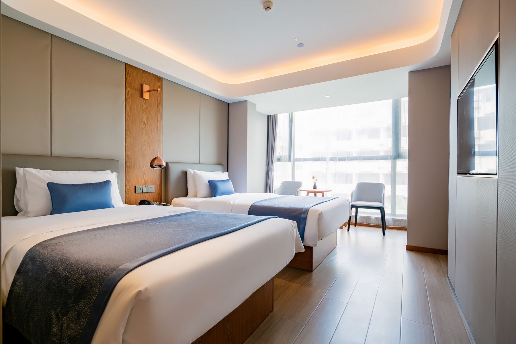 A hotel room with two beds, a wooden accent wall, a small table, and a large window.