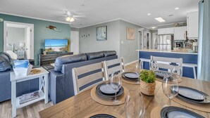 A modern living room with a sofa, a dining table set for four, and a kitchen in the background. - Beach Bella-Sleeps 8, 3 min Walk to Beach (Destin)