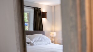 A bedroom with a bed, a nightstand, a lamp, and a window with curtains. - The New Inn (Cirencester)