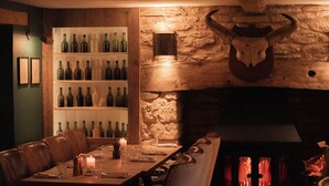 A dimly lit restaurant with a long wooden table set for dining, a stone wall with a bull's head, and shelves of wine bottles. - The New Inn (Cirencester)