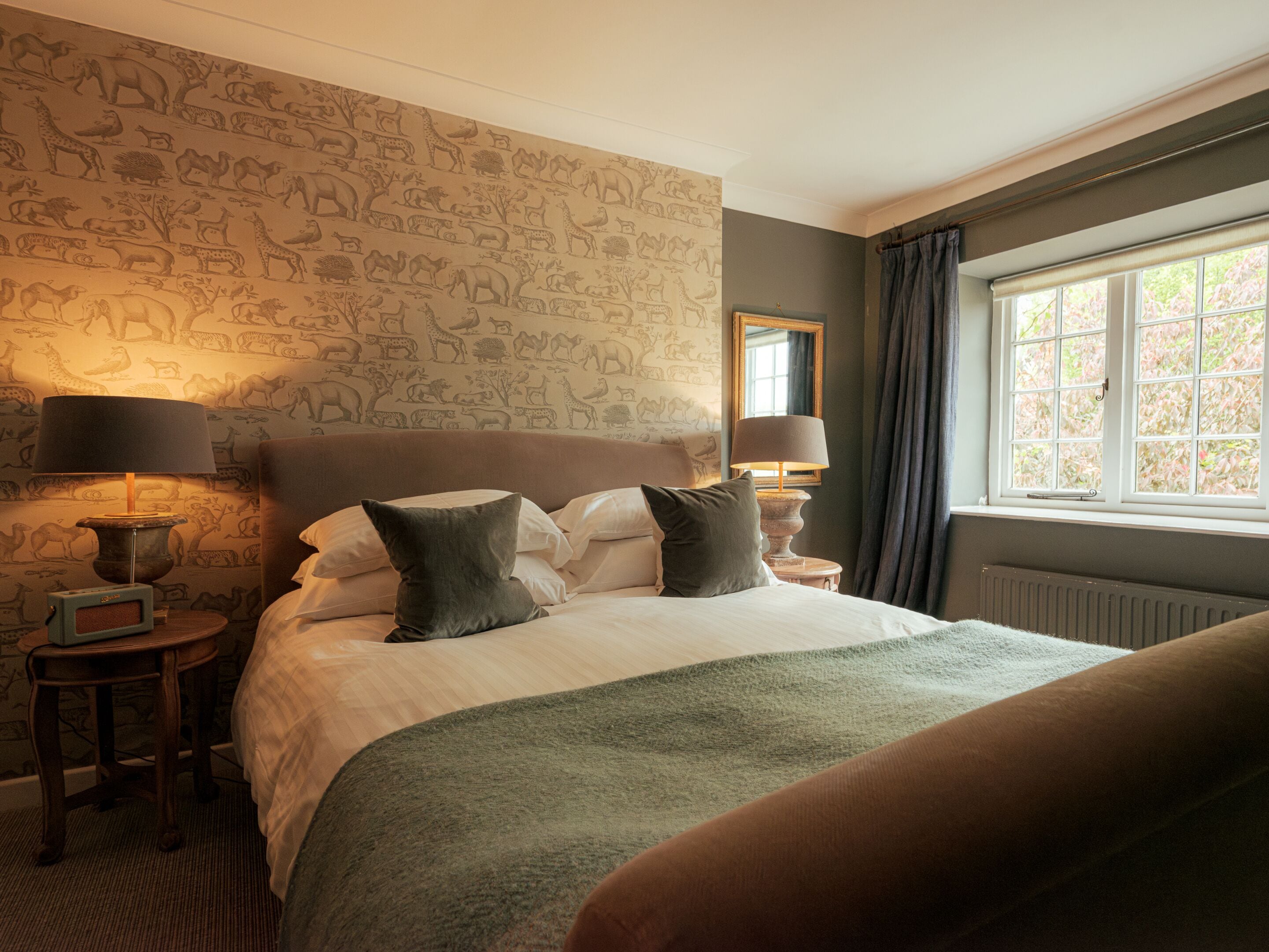 A bedroom with a large bed, two bedside tables with lamps, a window with curtains, and a wall with animal wallpaper.