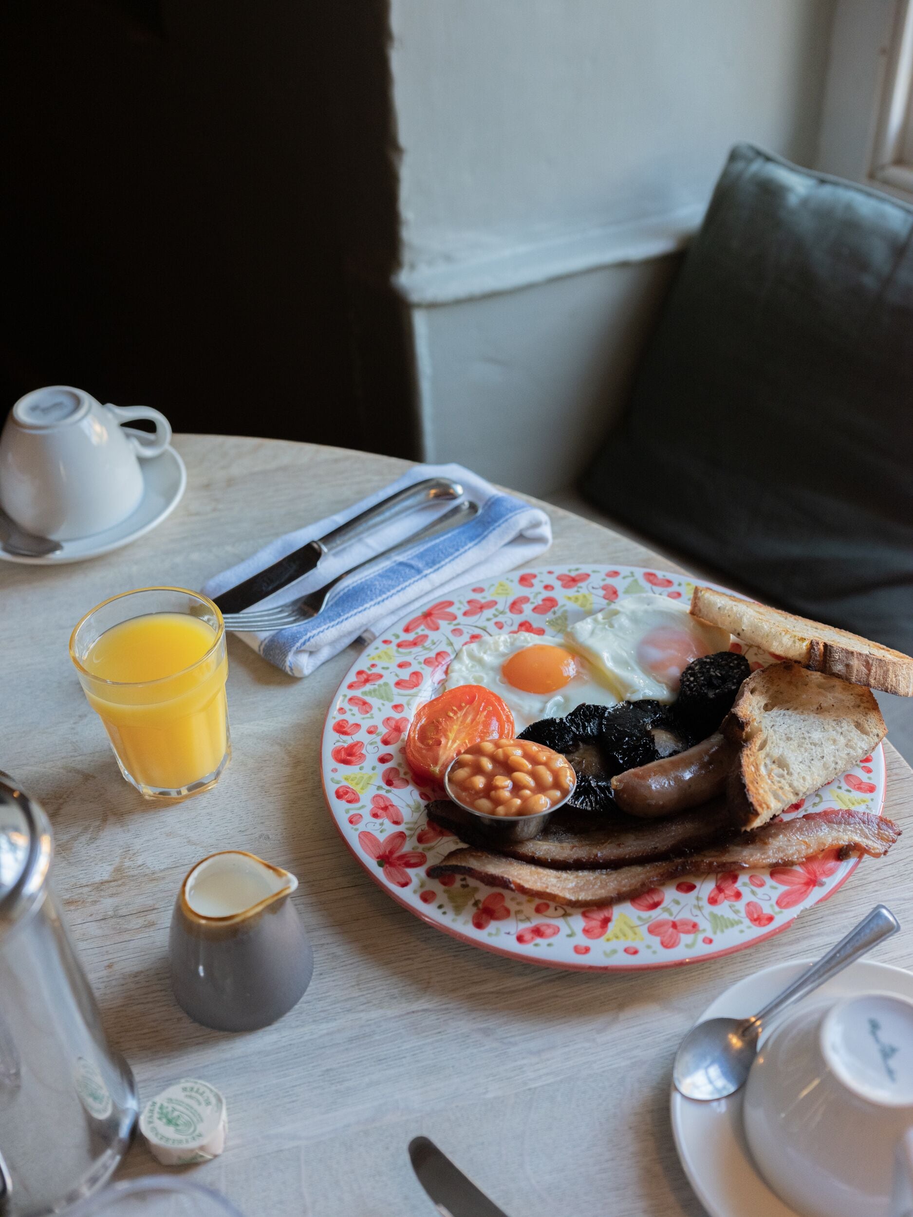 Una mesa de desayuno con un plato de huevos, salchichas, tostadas y frijoles, un vaso de jugo de naranja y una taza de té.
